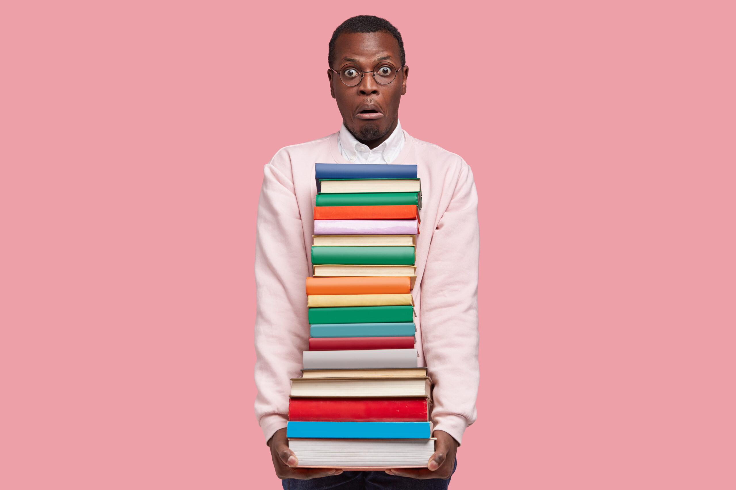 Stupefied young Afro American man with big pile of books, dressed in casual sweater, has surprised facial expression